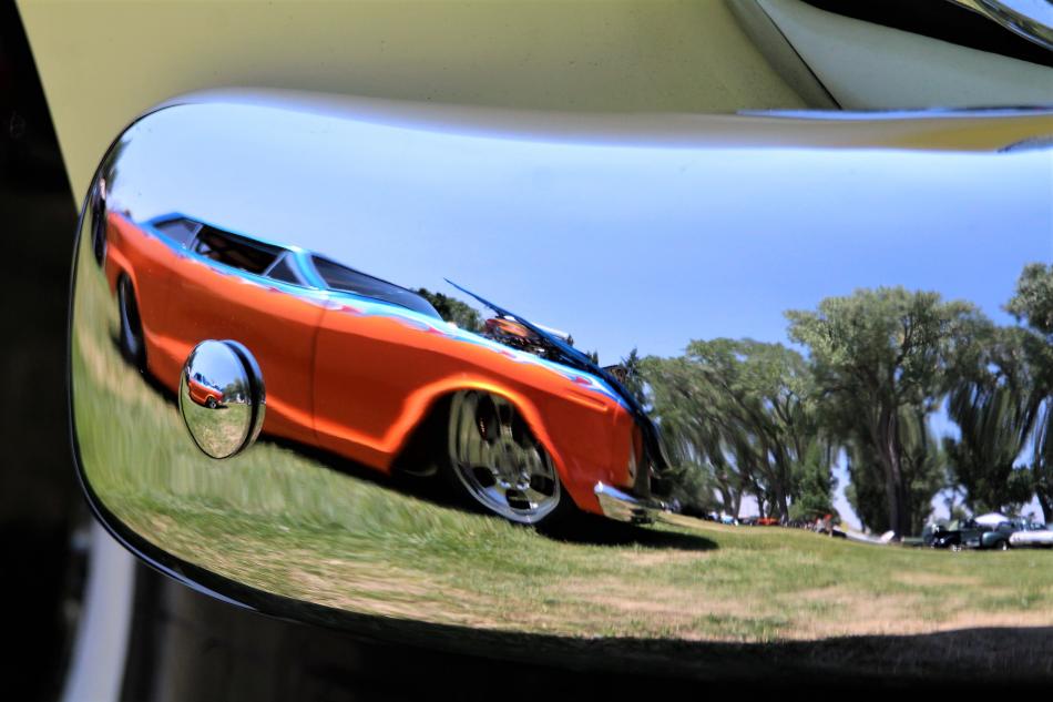 A custom street rod is reflected in the bumper of a neighboring classic car.