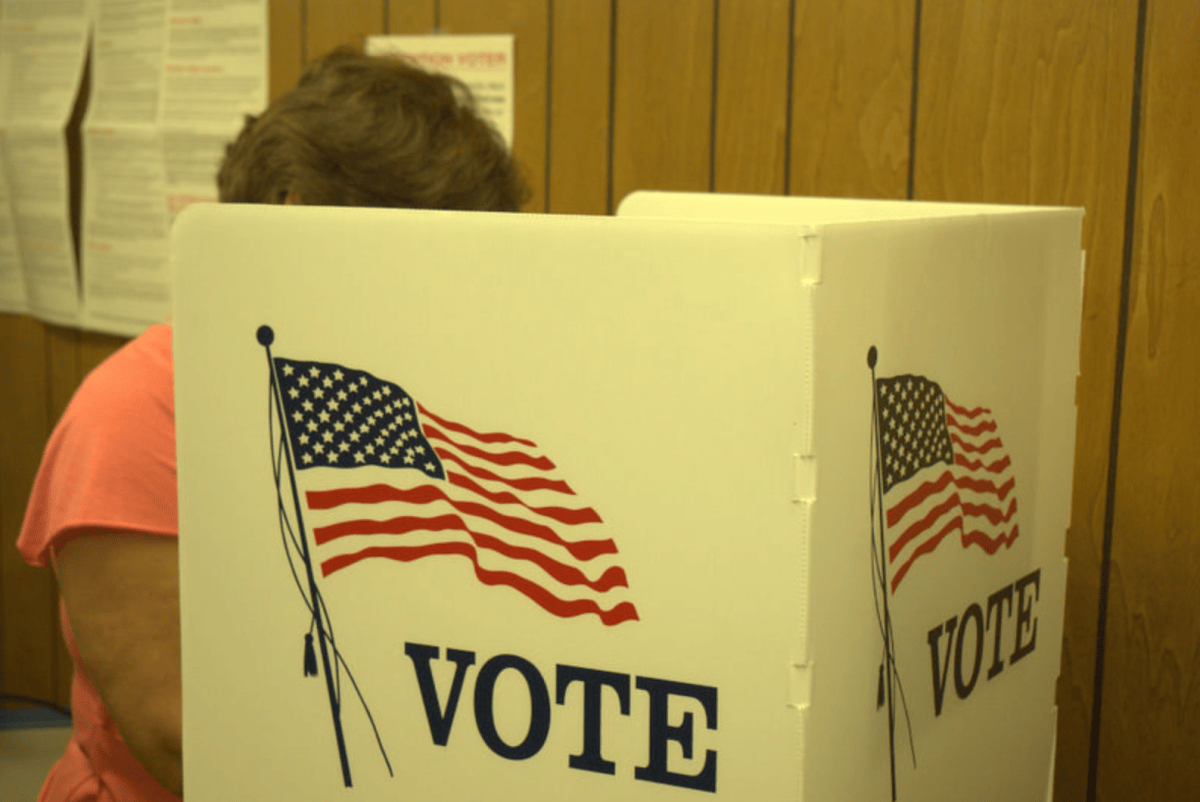 A voter leans into a poll box that says VOTE on the outside, their face is obscured