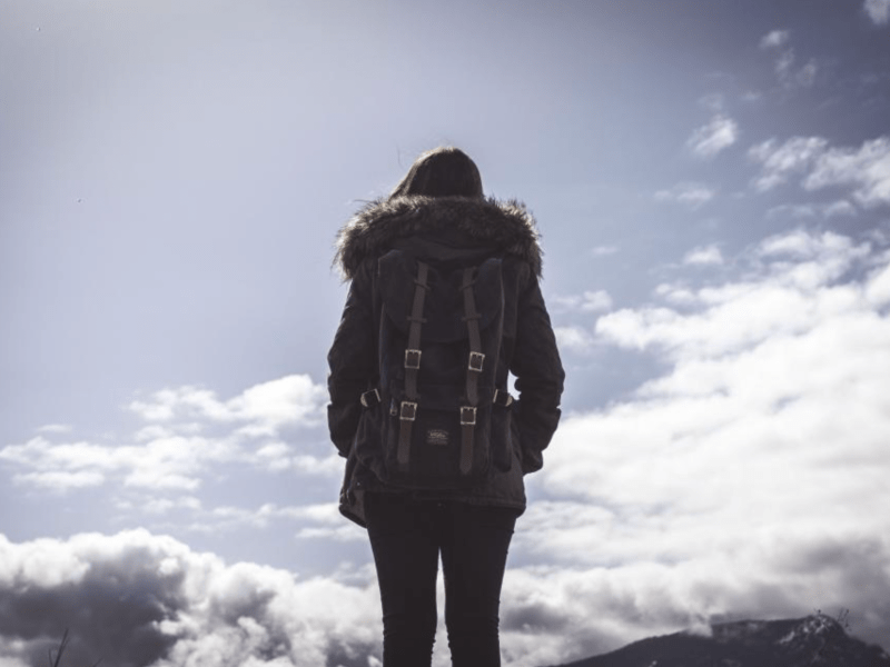 A woman with a backpack shown from behind looking at the sky where storm clouds are darkening in the distance.