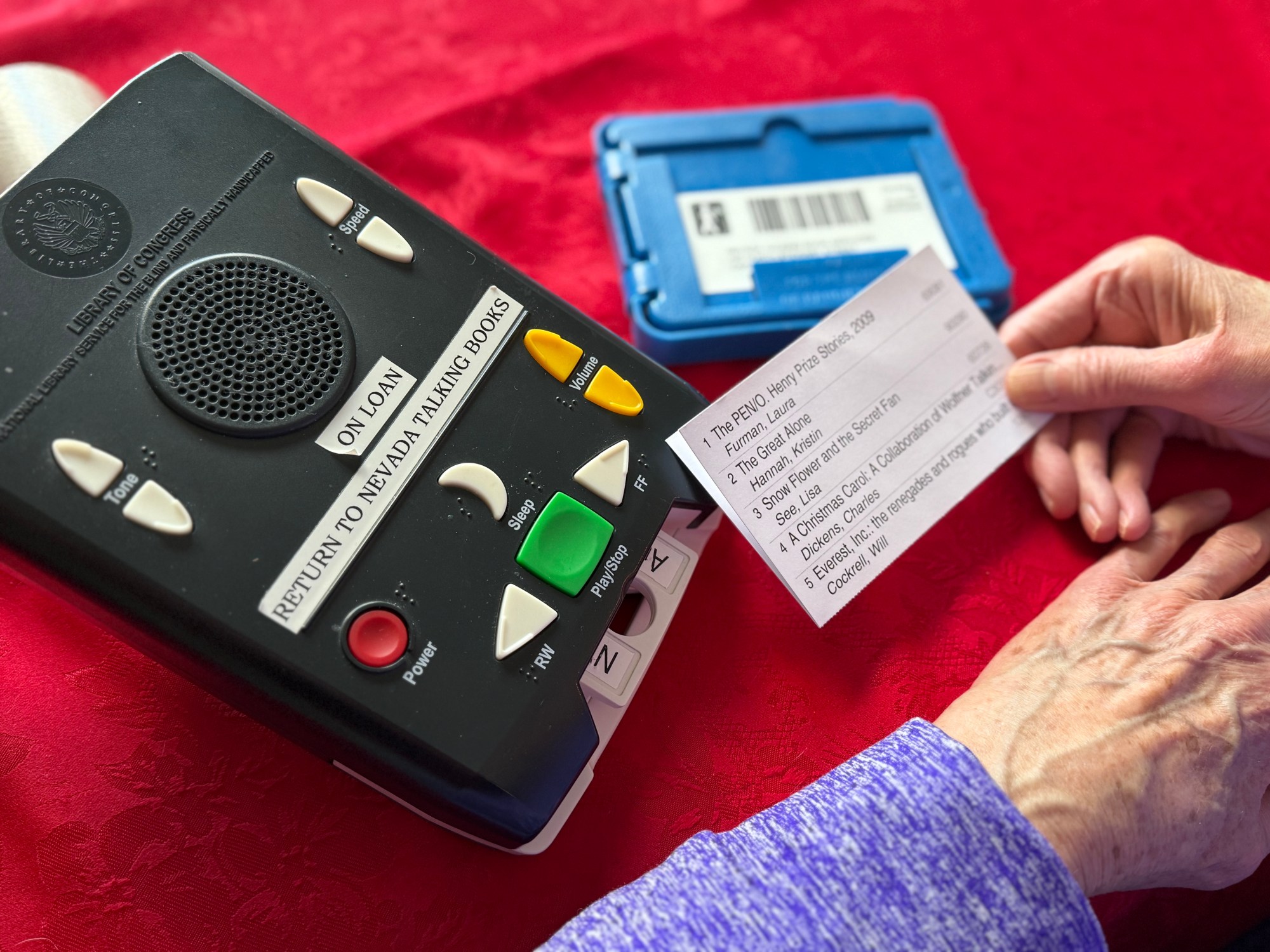 A tape player labeled Talking Book is on a red tablecloth, and a patron reads a card with the titles of audiobooks.