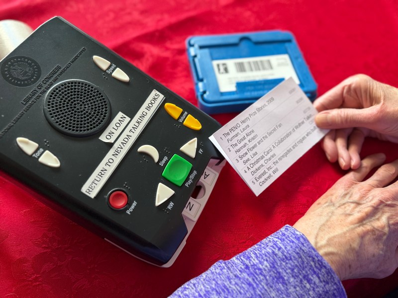 A tape player labeled Talking Book is on a red tablecloth, and a patron reads a card with the titles of audiobooks.